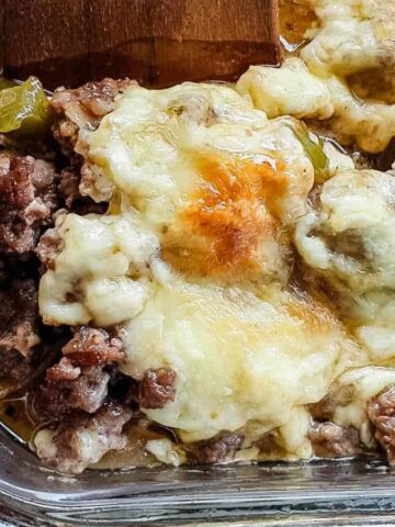 Close-up of a baked dish in a glass container, containing browned beef pieces, melted cheese, and green peppers. Part of the dish is resting on a wooden surface, and it sits on a wire rack.