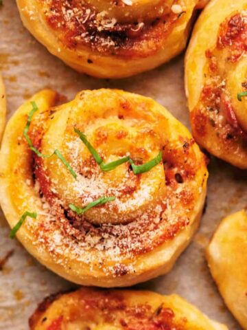 Close-up of golden-brown pizza rolls on a baking sheet, topped with melted cheese, tomato sauce, and sprinkled with fresh basil. Some rolls are garnished with grated cheese and herbs.