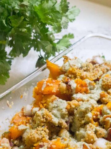 A glass dish contains a mixed salad with chunks of orange squash, crumbled toppings, and a green dressing. Fresh cilantro is on the table beside the dish.