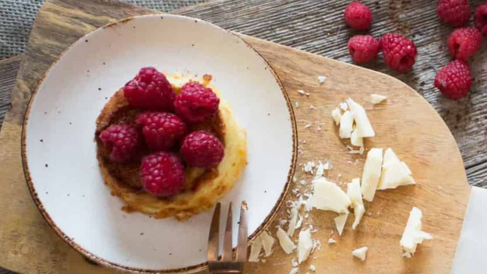A dessert placed on a white plate with a brown rim sits on a wooden board. The dessert is topped with raspberries. Shaved white chocolate pieces are scattered on the board. A fork rests beside the plate.
