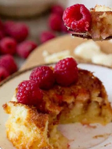 A fork holds a piece of cake above a white plate with a partially eaten slice of cake topped with raspberries. More raspberries and crumbs are scattered on a wooden board in the background.