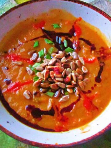 A bowl of pumpkin soup topped with sunflower seeds, drizzles of balsamic glaze, and chopped herbs. A spoon is placed on a green checkered tablecloth beside a slice of brown bread.