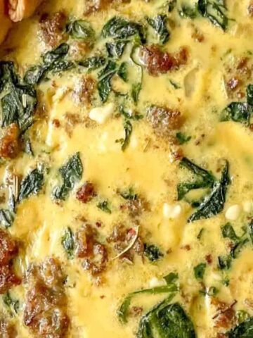 Close-up of a baked quiche in a white dish. The quiche has a golden crust and is filled with a mixture of creamy eggs, green spinach, and browned sausage. The dish is placed on a red cloth.