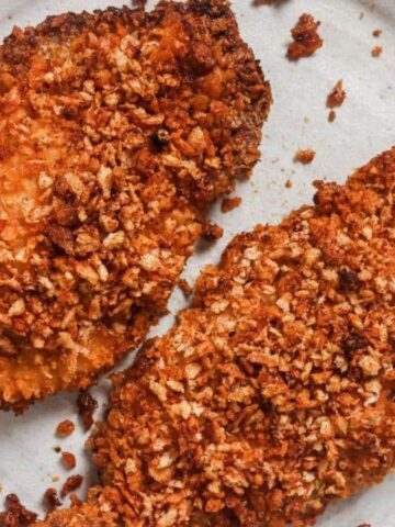 A white plate holds two pieces of crispy, breaded chicken. The chicken is golden brown with a textured, crunchy coating. Some small crumbs are scattered around the plate.