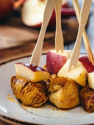A plate features skewered cubes of cooked meat and red apple slices on wooden sticks. The skewers are upright, and the plate rests on a wooden surface with blurred background items, including a bowl and a jar.