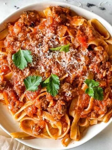 A white bowl filled with pasta topped with a rich tomato and meat sauce, garnished with fresh parsley and grated cheese. A fork and napkin are placed beside the bowl on a marble surface. A block of cheese is visible in the corner.