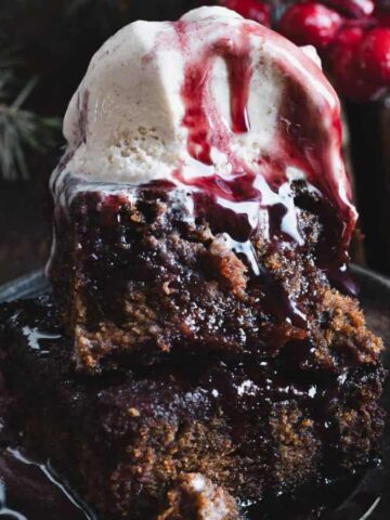 A slice of dark cake with a scoop of vanilla ice cream on top, drizzled with berry sauce, sits on a plate. Red berries and evergreen branches are in the background on a wooden surface.