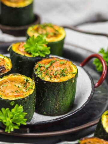 A plate of stuffed zucchini cups garnished with parsley is displayed. The zucchini is cut into thick slices and filled with a baked mixture. The presentation is on stacked round plates with red handles against a soft fabric background.
