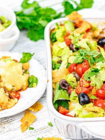 A casserole dish filled with a layered taco salad featuring lettuce, tomatoes, olives, tortilla chips, and a ground meat mixture. Next to it is a small plate with a serving of the salad and a fork. Fresh cilantro is in the background.