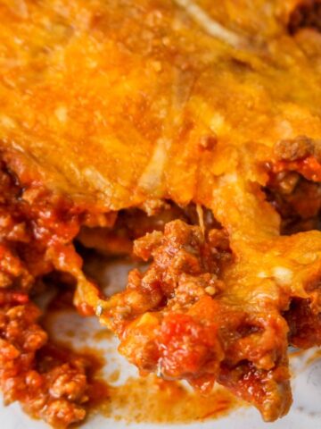 A close-up of a fork lifting a portion of lasagna from a plate. The lasagna features layers of melted cheese and tomato-based meat sauce. The dish sits on a white plate with a subtle decorative pattern.