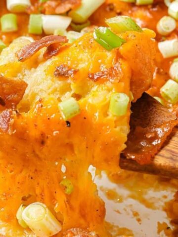 Close-up of a cheesy baked dish with a wooden spoon lifting a portion. The dish is topped with melted cheese, crispy bacon, and chopped green onions. The edge of a white baking dish is visible around the food.