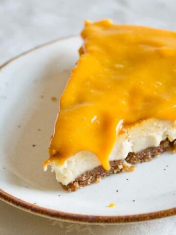 A slice of pie on a white plate with a brown rim. The pie has a crumbly base, a creamy filling, and is topped with a glossy orange layer. A fork is placed beside the plate on a white tablecloth.
