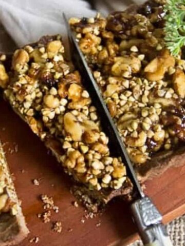 A rectangular nut tart on a wooden board is partially sliced with a knife. The tart is topped with various nuts, including walnuts and possible seeds, and a sprig of green leaves. A piece of tart is separated from the main portion.