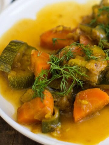 A white plate contains a dish of stewed vegetables, including chunks of carrots and zucchini, topped with fresh dill. The vegetables are covered in a yellow-orange sauce. A fork rests on the plate's edge.
