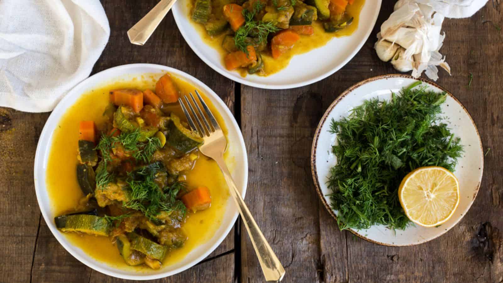 A white plate contains a dish of stewed vegetables, including chunks of carrots and zucchini, topped with fresh dill. The vegetables are covered in a yellow-orange sauce. A fork rests on the plate's edge.
