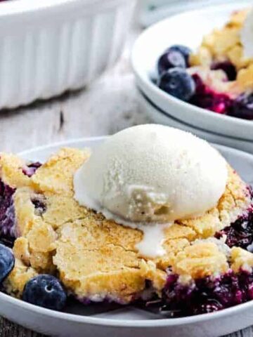 A dessert plate with a portion of blueberry cobbler topped with a scoop of vanilla ice cream. The cobbler features a golden crust and is surrounded by fresh blueberries. Another plate with the same dessert is visible in the background.