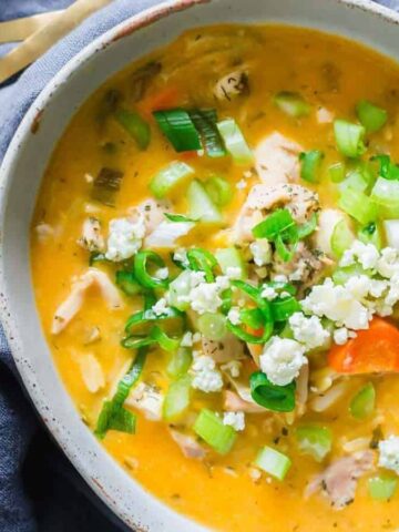 A bowl of hearty soup with orange broth, containing chunks of chicken, sliced carrots, and chopped celery. Topped with chopped green onions and crumbled cheese. Two golden spoons rest beside it on a dark napkin.