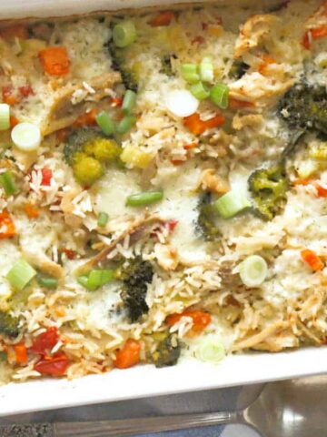 A baked casserole in a rectangular white dish contains rice, broccoli, red bell peppers, sliced green onions, and melted cheese. A spoon and fork are placed beside the dish on a striped cloth.