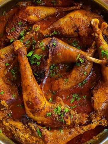 A pan filled with cooked chicken legs in a rich, red sauce garnished with herbs showcases a low-effort recipe that tastes like you've spent hours in the kitchen. Next to it, a bowl of white rice and a small dish of spices sit on a wooden table, with a knife resting on a beige cloth nearby.