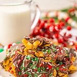 A plate of chocolate-covered cornflake brittle clusters is artfully decorated with red, white, and green sprinkles. A glass of milk stands in the background, surrounded by festive red and green decorations.