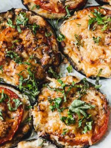 A white oval plate with baked eggplant slices topped with melted cheese, tomato, and chopped herbs. The dish is placed on a wooden table partially covered by a dark cloth.