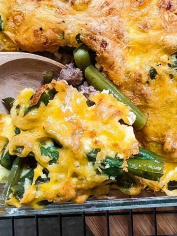 A glass baking dish holds a baked casserole with a melted cheese topping. Visible ingredients include green beans, leafy greens, and ground meat. A wooden spoon is scooping a portion from the dish.