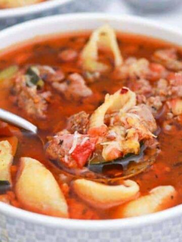 A bowl of hearty soup containing ground meat, shell pasta, diced tomatoes, and sliced zucchini in a tomato-based broth. A spoon rests in the bowl. In the background, a tomato and celery stalk are partially visible.