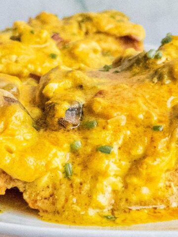 A plate of baked chicken breast covered with a melted cheese topping, mushrooms, and bits of ham. The dish is garnished with chopped herbs.