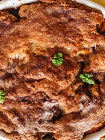 A baked apple cobbler with a golden-brown crust is displayed in a round dish. The surface is sprinkled with sugar and adorned with a few small green leaves. Slices of fresh peaches and green leaves surround the dish on a dark background.