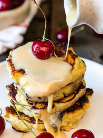 A stack of pancakes topped with cherries is on a white plate. Syrup is being poured over the pancakes from a jug. More cherries and a folded cloth are visible in the blurred background. The setting is a rustic wooden surface.