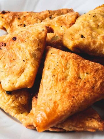 A white plate filled with golden brown, triangular samosas sits on a marble surface, crafted to perfection using Air Fryer techniques. The samosas are sprinkled with black pepper, while a sprig of fresh cilantro peeks from the background. Indulge in these delights and fry without guilt!