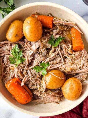 A bowl of shredded meat is served with whole potatoes and large carrot pieces, garnished with parsley. The dish is on a marble surface, next to a bunch of fresh parsley, a small bowl of fennel seeds, and a red cloth with a fork.