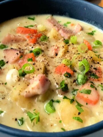 A blue bowl filled with creamy soup containing chunks of vegetables and meat, garnished with chopped green onions and herbs, makes for a perfect lazy weekend meal. The dish rests on a white plate surrounded by round crackers, truly begging for seconds.
