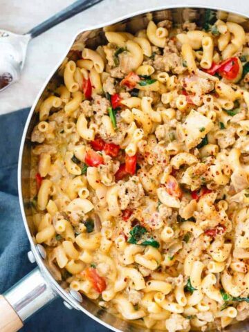 A pot filled with cooked macaroni mixed with ground meat, chopped tomatoes, and spinach, garnished with a sprinkle of spices. Fresh basil leaves are placed beside the pot on a dark cloth.