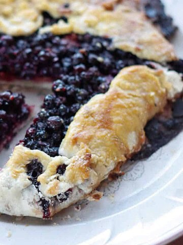 A rustic blueberry galette on a white plate. The pastry is golden brown and folded over the edges of the blueberry filling. One slice has been removed, revealing the juicy interior. The plate is on a wooden surface.