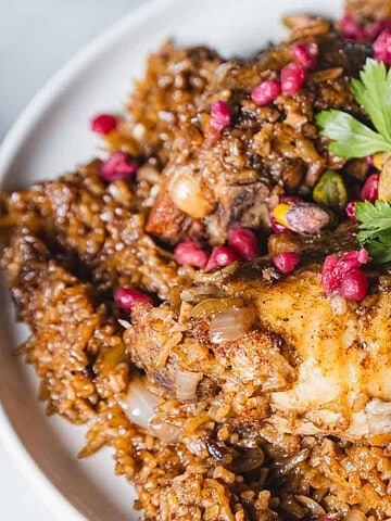 A plate of rice topped with cooked chicken pieces and garnished with fresh parsley leaves. The dish is sprinkled with red pomegranate seeds and pistachios, providing a colorful contrast to the brown rice and chicken.