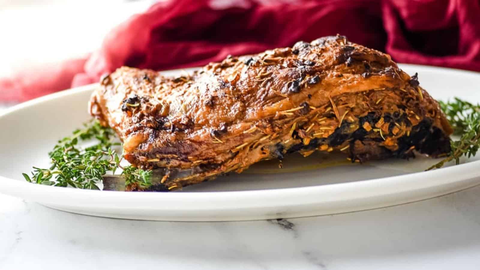 A seasoned, cooked lamb shoulder rests on a white plate garnished with sprigs of fresh thyme&mdash;a perfect centerpiece for those who cherish warm, filling meals. A red cloth draped in the background adds a splash of color to the setting. The meat appears tender and well-roasted.