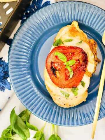 A cooked chicken breast topped with melted cheese, sliced tomato, and fresh basil on a blue plate invites you to try tonight. A gold fork is placed beside it. In the background, there is a grilled cooking tray, a patterned blue napkin, and a rolled white napkin.