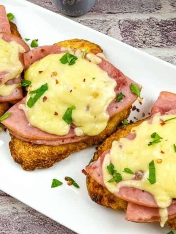 On a white rectangular plate, three servings of toasted bread are topped with slices of ham and melted cheese, perfect for laziest weeknights. The dish is garnished with chopped parsley and a sprinkle of red pepper flakes. A bowl of salad is partially visible in the background.