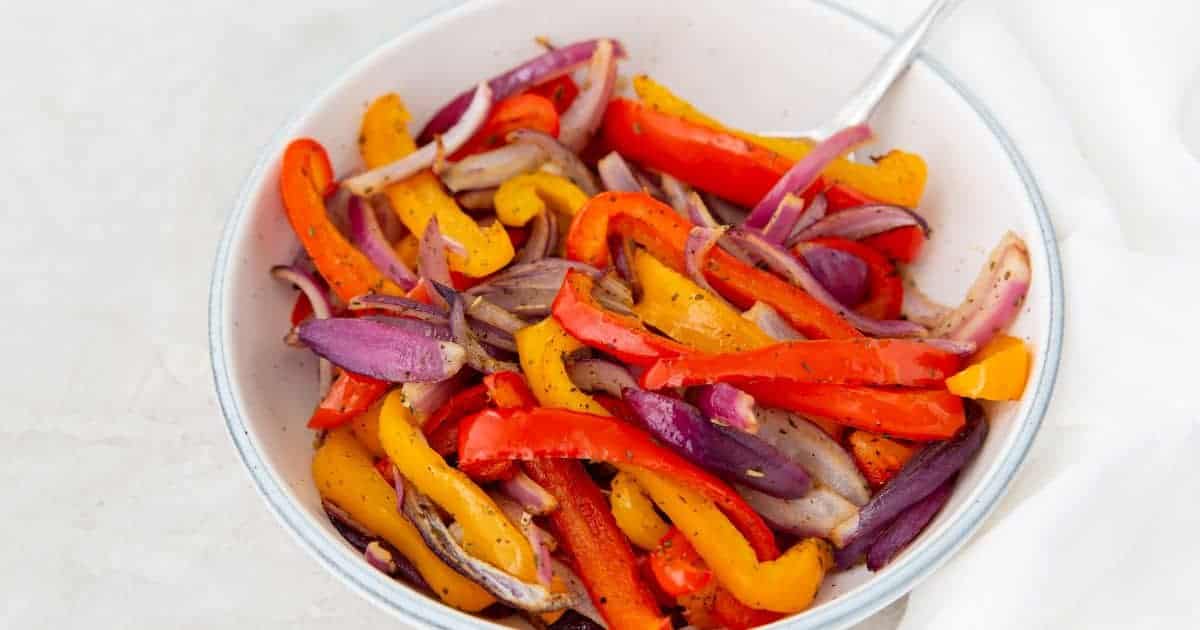 A white bowl brimming with must-try, air-fried red, yellow, and orange bell peppers alongside red onions. Seasoned perfectly with black pepper, these roasted veggies promise effortless cooking. A spoon rests invitingly in the bowl against a light-colored surface.