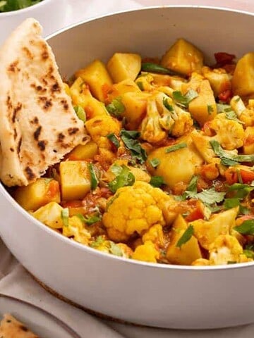 A white pan brims with a vibrant yellow curry, featuring tender potatoes and cauliflower, all garnished with fresh cilantro. A piece of naan rests invitingly on the side, perfect for indulgent weekend meals spent unwinding. Plates and a napkin subtly suggest a shared feast in the background.