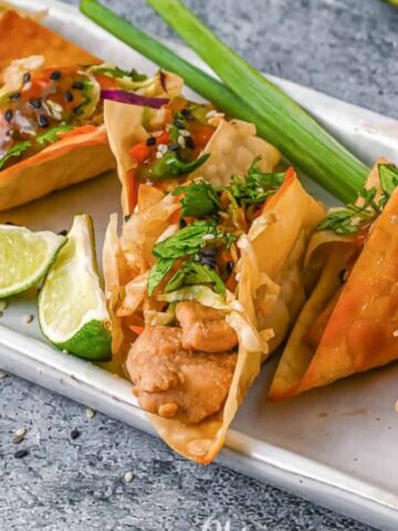 A rectangular plate features golden-brown crispy taco shells filled with chicken, shredded vegetables, and garnished with cilantro and sesame seeds. Perfect for sharing cherished meals with loved ones, lime wedges and green onions are placed beside the tacos on a textured gray surface.