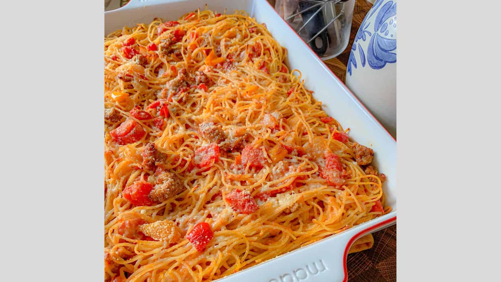 A budget-friendly baked spaghetti dish with red sauce, chunks of tomato, and grated cheese on top is served in a rectangular white dish. A blue and white patterned mug is visible to the right, perfect for cozy dinners that are easy on your wallet.