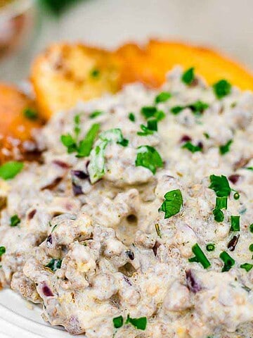 A white plate with a serving of creamy sausage gravy topped with chopped green herbs, accompanied by several slices of toasted bread. The background includes a blurred view of additional herbs and a small bowl.
