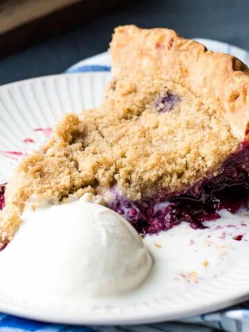 A slice of berry crumble pie with a golden-brown topping is served on a white plate. A scoop of vanilla ice cream is melting beside it. The plate rests on a blue and white striped cloth.