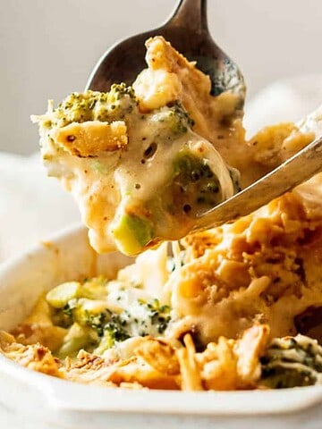 A spoonful of creamy broccoli and chicken casserole being lifted from a white dish. This comforting treat, topped with browned, crispy bits, is perfect for keeping the winter blues at bay. A blurred background reveals a white tablecloth and a hint of greenery.