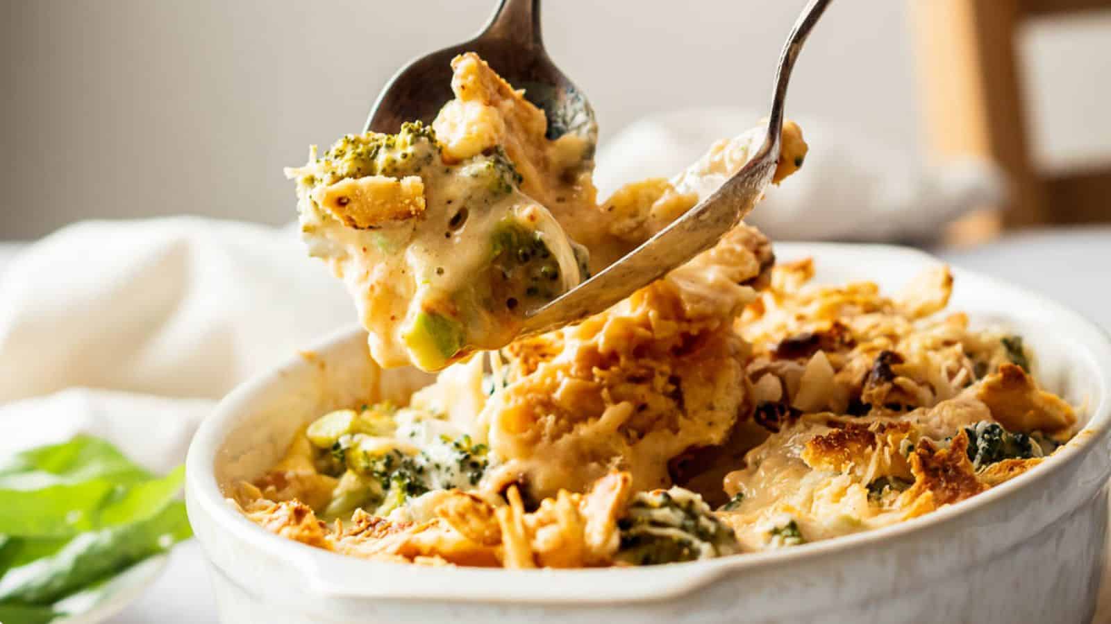 A spoonful of creamy broccoli and chicken casserole being lifted from a white dish. This comforting treat, topped with browned, crispy bits, is perfect for keeping the winter blues at bay. A blurred background reveals a white tablecloth and a hint of greenery.