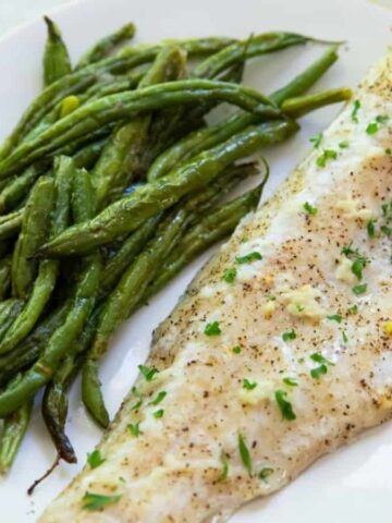A white plate features a seasoned white fish fillet alongside tender green beans, both artfully garnished with chopped herbs. This entree is sure to be a hit among those seeking fresh and flavorful recipes.