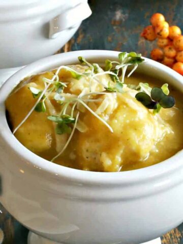 A white ceramic bowl contains creamy yellow soup garnished with greens, offering a taste of winter comfort foods. Nearby are golden spoons, a beige napkin, and an arrangement of small orange berries on a blue wooden surface—perfect for those cozy evenings when you want to make tonight special.