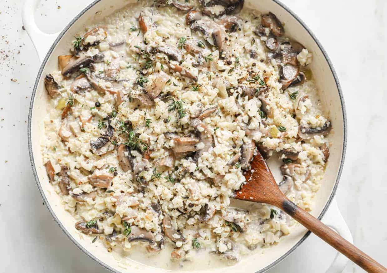 A pan of creamy risotto with mushrooms and herbs is being stirred with a wooden spoon. The dish contains visible pieces of mushrooms and is garnished with fresh thyme. The risotto appears rich and well-blended.
